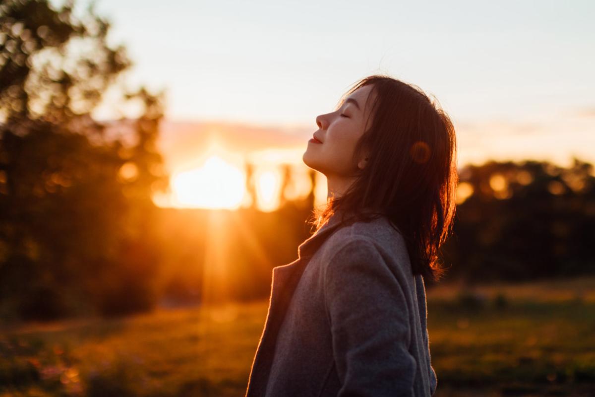 A woman standing in a field, smiling and inhaling the air, as the sun sets behind her.