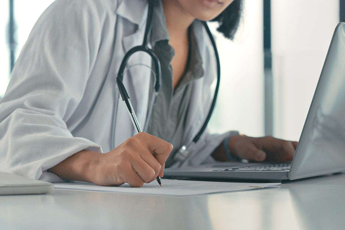 Physician sitting at a desk writing down notes while working on a laptop