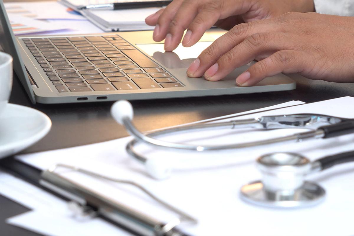 Hands typing on laptop with stethoscope and clipboard nearby